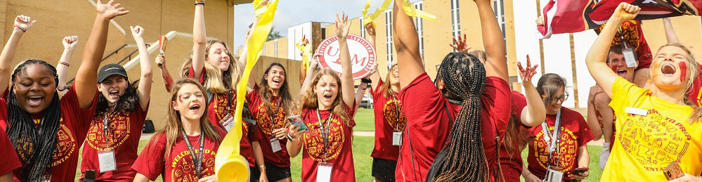 photo of about a dozen  students wearing matching marron and gold shirts cheer with their arms raised. With streamers and flags flying, the students exude high energy on a grassy lawn with yellow brick buildings in the background.